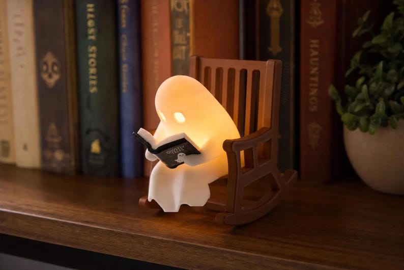 Three-quarter view of a softly glowing white reading ghost holding an open book in its brown rocking chair, displayed on a bookshelf beside a small potted plant.