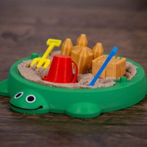 May include: A green turtle-shaped sandbox toy with a smiling face. Inside, there's sand, a yellow rake, a red bucket, a blue stick, and miniature sandcastles. The toy is designed for children's play.