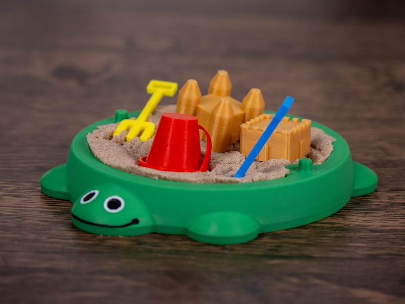 May include: A green turtle-shaped sandbox toy with a smiling face. Inside, there's sand, a yellow rake, a red bucket, a blue stick, and miniature sandcastles. The toy is designed for children's play.