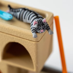 May include: A miniature cat tree with a gray and black striped cat figurine and a blue fish toy. The cat is lying down on the top level of the wooden cat tree. An orange support beam is visible.