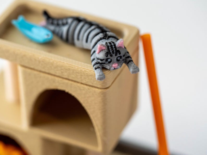May include: A miniature cat tree with a gray and black striped cat figurine and a blue fish toy. The cat is lying down on the top level of the wooden cat tree. An orange support beam is visible.