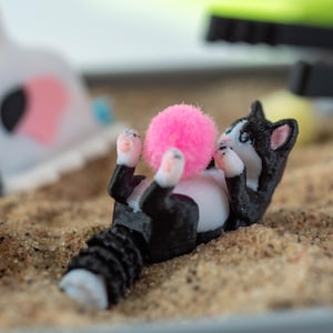 May include: A 3D-printed black and white cat figurine is lying on its back, paws up, playing with a bright pink pom-pom. The cat is in a sandbox with other colorful toys in the background.