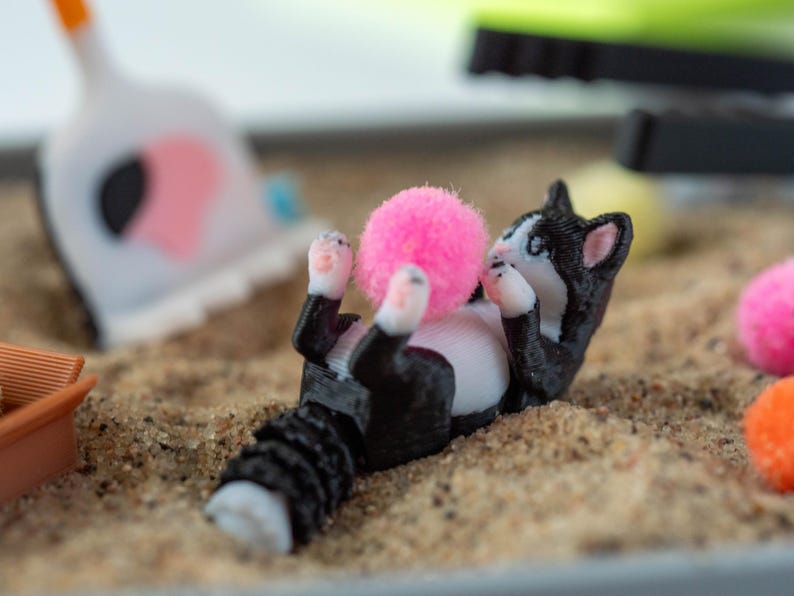May include: A 3D-printed black and white cat figurine is lying on its back, paws up, playing with a bright pink pom-pom. The cat is in a sandbox with other colorful toys in the background.
