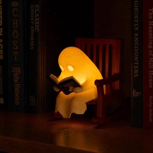 Reading ghost glowing yellow in a brown miniature rocking chair between upright ghost-story and horror books on a dark wooden shelf.