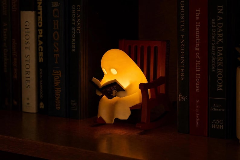 Reading ghost glowing yellow in a brown miniature rocking chair between upright ghost-story and horror books on a dark wooden shelf.