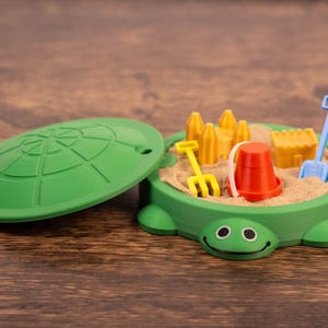 May include: A green turtle-shaped sandbox toy with a removable lid. Inside, there's sand and miniature toys: a yellow rake, a red bucket, a blue shovel, and yellow sandcastle molds. The toy is on a wooden surface.