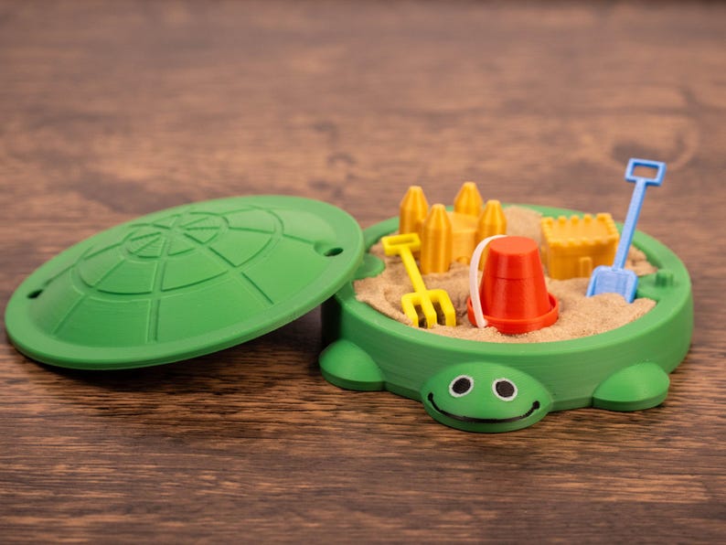 May include: A green turtle-shaped sandbox toy with a removable lid. Inside, there's sand and miniature toys: a yellow rake, a red bucket, a blue shovel, and yellow sandcastle molds. The toy is on a wooden surface.