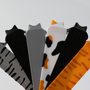 May include: A selection of cat-shaped door stoppers in different colours and patterns. The stoppers include grey with black stripes, solid grey, black, white with black and orange spots, solid black, and orange with stripes.