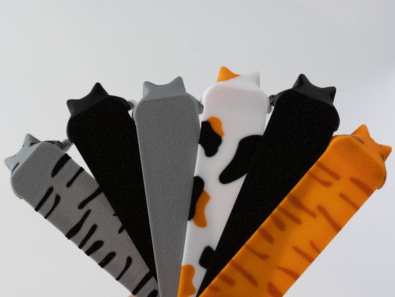 May include: A selection of cat-shaped door stoppers in different colours and patterns. The stoppers include grey with black stripes, solid grey, black, white with black and orange spots, solid black, and orange with stripes.