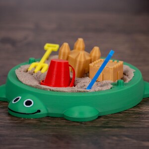 May include: A green turtle-shaped sandbox toy filled with sand and miniature accessories. Includes a red bucket, yellow rake, blue stick, and sandcastle molds. The toy is designed for children's play and creative activities.
