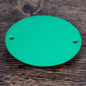 May include: A bright green, circular object with two small holes, possibly a component or part. The object is set against a dark wood grain background, providing a contrast in color and texture.