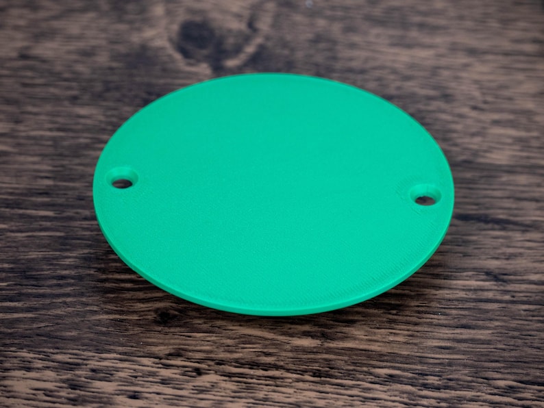 May include: A bright green, circular object with two small holes, possibly a component or part. The object is set against a dark wood grain background, providing a contrast in color and texture.