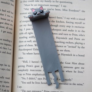 May include: A grey cat bookmark with a smiling face and pink details. The bookmark, made of a flat, ribbon-like material, is inserted in a book. The cat's paws and tail are visible at the bottom. The bookmark is a cute reading accessory.