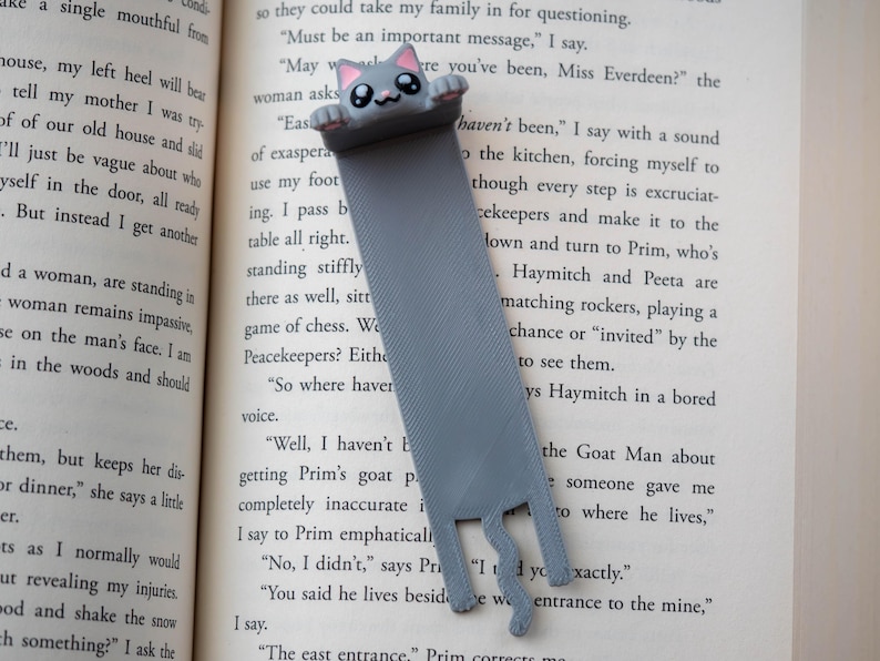 May include: A grey cat bookmark with a smiling face and pink details. The bookmark, made of a flat, ribbon-like material, is inserted in a book. The cat's paws and tail are visible at the bottom. The bookmark is a cute reading accessory.