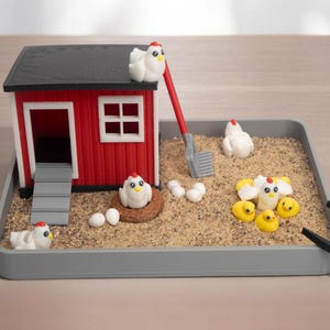 Puede incluir: Un gallinero en miniatura colocado en una bandeja gris llena de arena. El gallinero rojo y negro cuenta con una rampa, una pala roja y varios huevos blancos. Peque&ntilde;as figuras de gallinas blancas y pollitos amarillos decoran la arena.