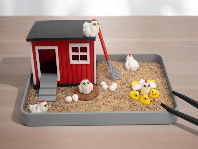 Puede incluir: Un gallinero en miniatura colocado en una bandeja gris llena de arena. El gallinero rojo y negro cuenta con una rampa, una pala roja y varios huevos blancos. Peque&ntilde;as figuras de gallinas blancas y pollitos amarillos decoran la arena.