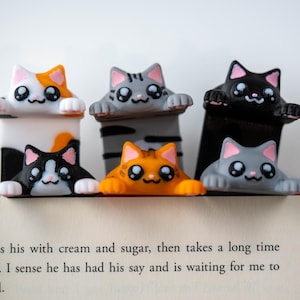 May include: Six cat-shaped bookmarks in different colours, including orange, grey, black, and calico. Each bookmark has a cute, cartoonish design with large eyes and paws. They are resting on the edge of an open book.
