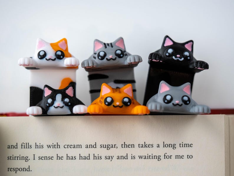 May include: Six cat-shaped bookmarks in different colours, including orange, grey, black, and calico. Each bookmark has a cute, cartoonish design with large eyes and paws. They are resting on the edge of an open book.