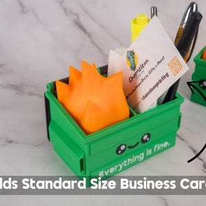 Puede incluir: Un organizador de escritorio en forma de cubo de basura impreso en 3D, de color verde y naranja, con una cara sonriente y el texto "Todo est&aacute; bien". El cubo de basura contiene tarjetas de visita, bol&iacute;grafos y un rotulador.