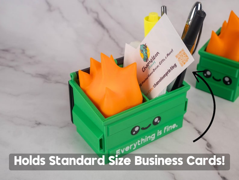 Puede incluir: Un organizador de escritorio en forma de cubo de basura impreso en 3D, de color verde y naranja, con una cara sonriente y el texto "Todo est&aacute; bien". El cubo de basura contiene tarjetas de visita, bol&iacute;grafos y un rotulador.