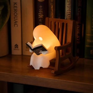 Warmly lit reading ghost in a brown rocking chair on a wooden bookshelf, with upright books behind it, a potted plant on the left and part of a white vase on the right.