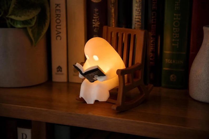 Warmly lit reading ghost in a brown rocking chair on a wooden bookshelf, with upright books behind it, a potted plant on the left and part of a white vase on the right.