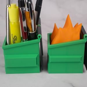 Puede incluir: Dos mini contenedores de basura de pl&aacute;stico verde, uno con un resaltador amarillo y varios bol&iacute;grafos en su interior, el otro con un objeto naranja en forma de llama en su interior.