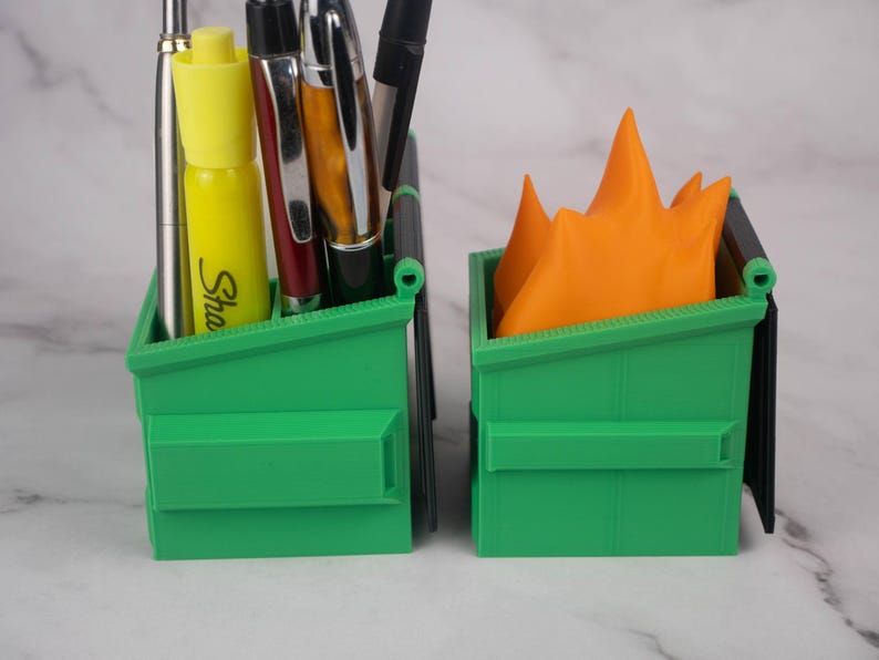 Puede incluir: Dos mini contenedores de basura de pl&aacute;stico verde, uno con un resaltador amarillo y varios bol&iacute;grafos en su interior, el otro con un objeto naranja en forma de llama en su interior.