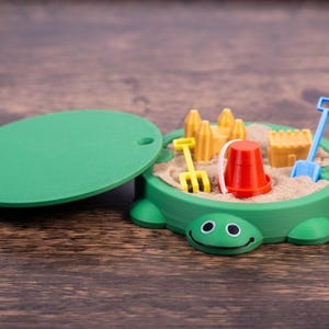 May include: A green turtle-shaped sandbox toy with a lid, filled with sand and miniature toys. Includes a red bucket, yellow rake, blue shovel, and yellow sandcastle molds. The lid is green and oval-shaped.
