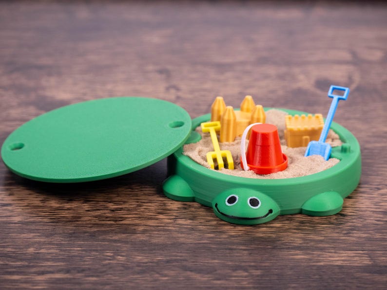 May include: A green turtle-shaped sandbox toy with a lid, filled with sand and miniature toys. Includes a red bucket, yellow rake, blue shovel, and yellow sandcastle molds. The lid is green and oval-shaped.