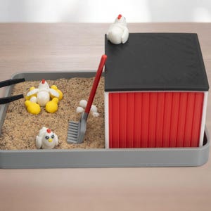 Puede incluir: Una bandeja sensorial en miniatura con tem&aacute;tica de granja que incluye una base rectangular gris llena de arena, un granero rojo y blanco con techo negro, tres figuras de pollos blancos con patas amarillas, un rastrillo rojo y pinzas negras.