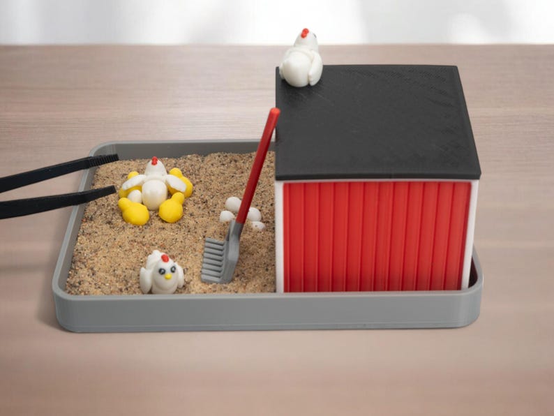 Puede incluir: Una bandeja sensorial en miniatura con tem&aacute;tica de granja que incluye una base rectangular gris llena de arena, un granero rojo y blanco con techo negro, tres figuras de pollos blancos con patas amarillas, un rastrillo rojo y pinzas negras.