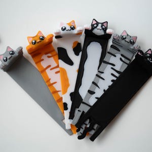 May include: Six cat-shaped bookmarks in various colours. The designs include a grey cat, an orange tabby, a calico, a black and white cat, a grey tabby, and a black cat. The cat heads are at the top, with the bookmark tails extending downwards.