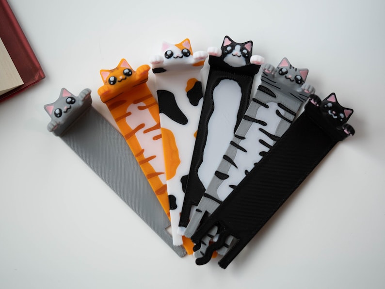 May include: Six cat-shaped bookmarks in various colours. The designs include a grey cat, an orange tabby, a calico, a black and white cat, a grey tabby, and a black cat. The cat heads are at the top, with the bookmark tails extending downwards.