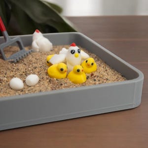 Puede incluir: Una bandeja rectangular gris llena de arena que contiene una gallina blanca en miniatura, tres pollitos amarillos peque&ntilde;os, dos huevos blancos y un rastrillo de pl&aacute;stico rojo y gris. La escena incluye una estructura roja parcial a la izquierda.