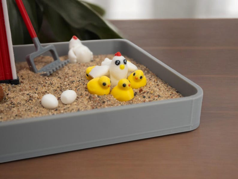 Puede incluir: Una bandeja rectangular gris llena de arena que contiene una gallina blanca en miniatura, tres pollitos amarillos peque&ntilde;os, dos huevos blancos y un rastrillo de pl&aacute;stico rojo y gris. La escena incluye una estructura roja parcial a la izquierda.