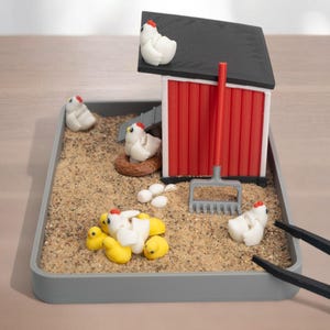 Puede incluir: Una escena de granja en miniatura en una bandeja rectangular gris llena de arena, que incluye un gallinero rojo y blanco con techo negro, varias gallinas blancas, pollitos amarillos, un nido marr&oacute;n, huevos blancos y un rastrillo gris.