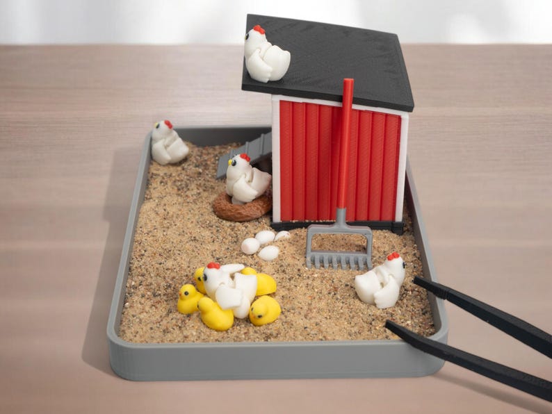 Puede incluir: Una escena de granja en miniatura en una bandeja rectangular gris llena de arena, que incluye un gallinero rojo y blanco con techo negro, varias gallinas blancas, pollitos amarillos, un nido marr&oacute;n, huevos blancos y un rastrillo gris.