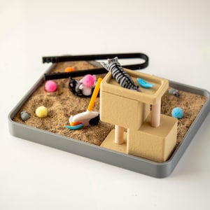 May include: A miniature cat-themed playset with a gray tray filled with sand. Includes a beige cat tower, toy cat, and various colorful toy balls. A black U-shaped tool and a toy golf club are also present.