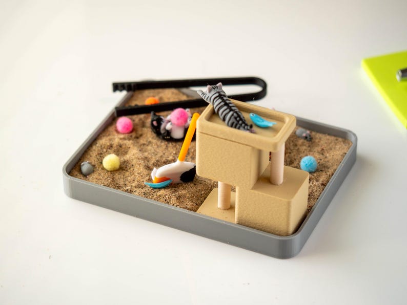 May include: A miniature cat-themed playset with a gray tray filled with sand. Includes a beige cat tower, toy cat, and various colorful toy balls. A black U-shaped tool and a toy golf club are also present.