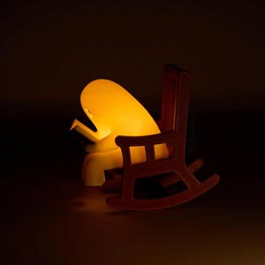 Side view of the glowing reading ghost leaning over its open book in a brown rocking chair with a tall slatted back, an armrest and curved rockers.