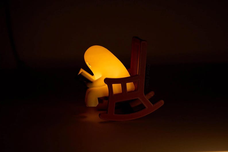 Side view of the glowing reading ghost leaning over its open book in a brown rocking chair with a tall slatted back, an armrest and curved rockers.