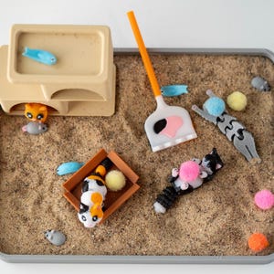 May include: A miniature cat-themed playset with a sand-like base. The set includes toy cats, fish, a scoop, and colorful pom-poms. A beige structure with a blue fish on top and a cat and mouse underneath is also present.