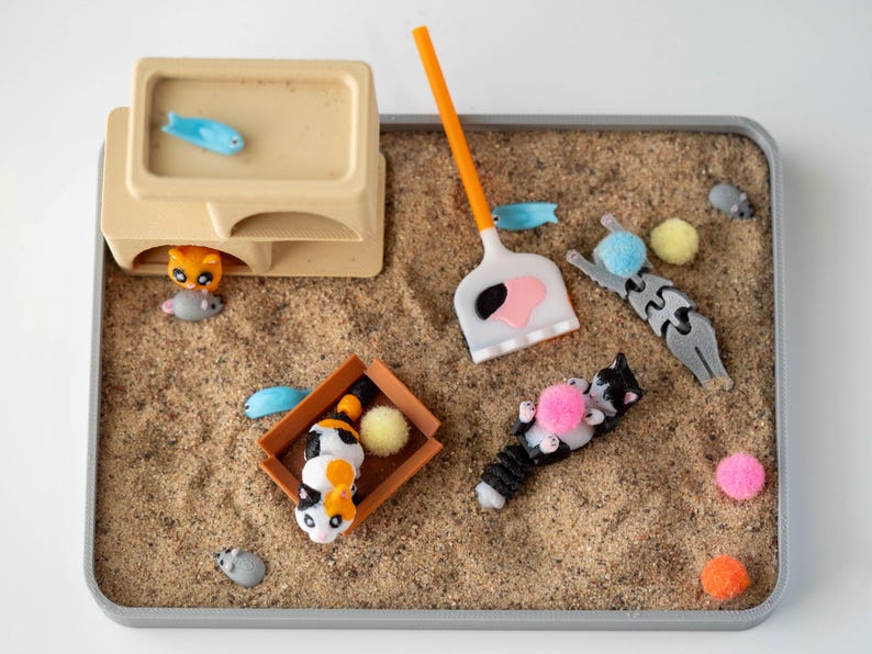 May include: A miniature cat-themed playset with a sand-like base. The set includes toy cats, fish, a scoop, and colorful pom-poms. A beige structure with a blue fish on top and a cat and mouse underneath is also present.