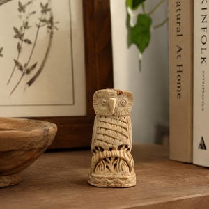 May include: A small, carved owl figurine in a light beige colour. The owl has detailed features, including carved feathers and facial details. The figurine is standing on a wooden surface, with a framed picture and books in the background.