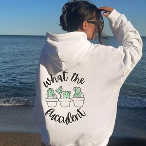 May include: White hoodie with a cactus graphic and the text "what the accident" in black script. The graphic features three potted cacti in shades of green. The hoodie has a hood and long sleeves.