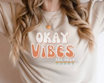 May include: A beige sweatshirt with the text "OKAY VIBES ARE OKAY" in a retro font. The text is in peach and white with a small design of triangles below.