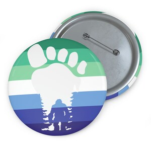 May include: A round button with a green, white, and blue striped background. A white footprint is in the center of the button, with a silhouette of a Bigfoot standing in the trees behind it.