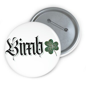 May include: A white button with a black and white graphic of the word "Birdy" in a gothic font. A green four-leaf clover with a leopard print pattern is to the right of the word.