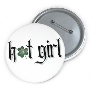 May include: A white button with a black gothic font that reads "hot girl" with a green four-leaf clover with a leopard print design in the center.
